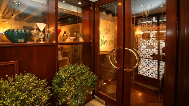Une entrée avec un cadre en bois et des portes en verre, un pot de fleurs et une fenêtre décorative en verre portant l’inscription « Cu’ Del Bryone ».