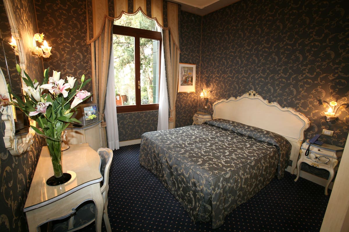 A hotel room with a bed, a desk, a chair, a vase with flowers, and a window with curtains.