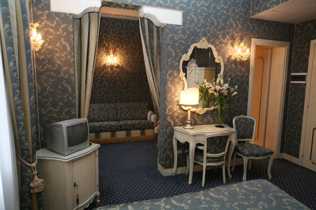 A room with a patterned wallpaper, a television on a cabinet, a desk with a lamp and flowers, a chair, and a mirror.