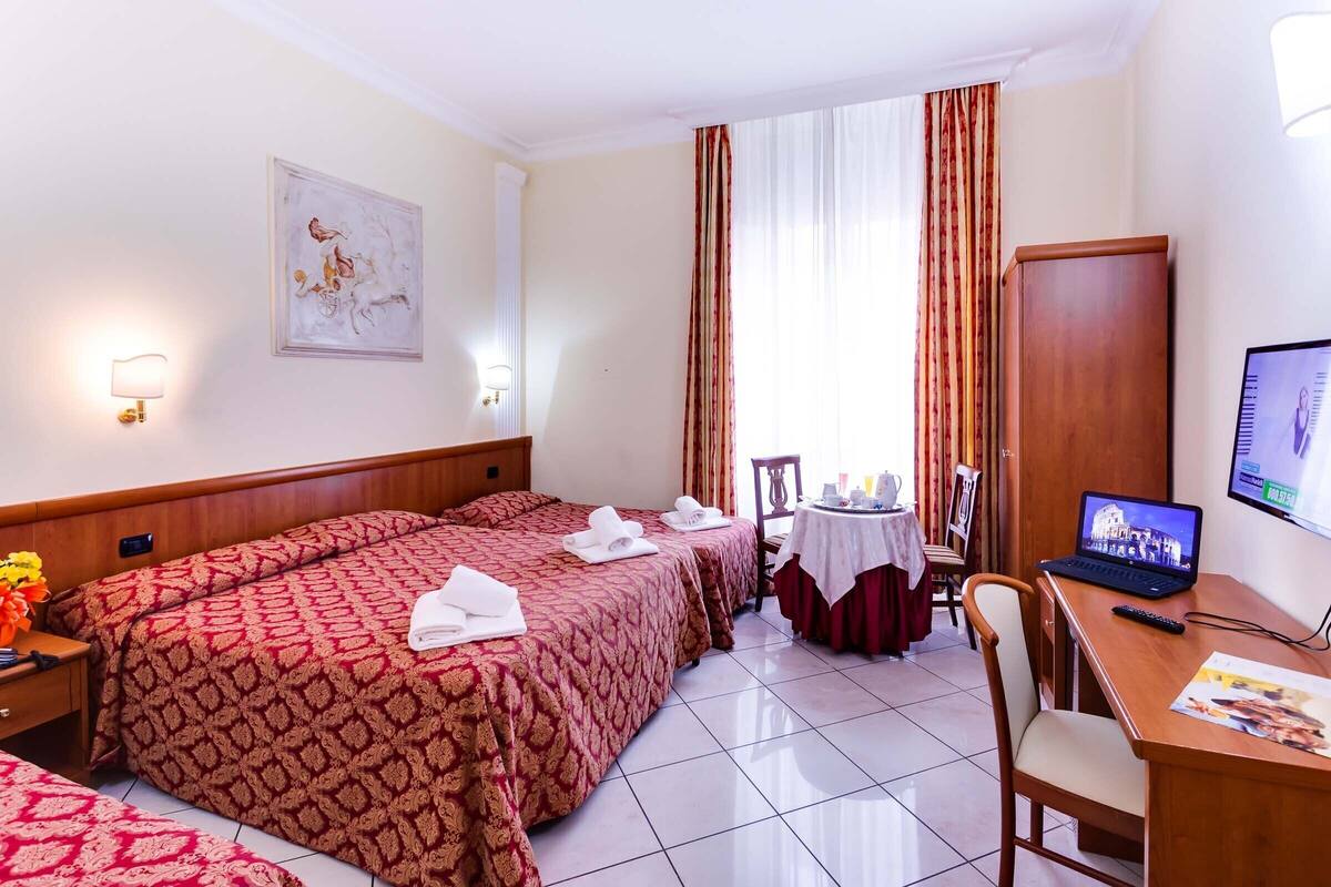 A hotel room with two beds, a desk with a laptop, a TV, and a window with curtains.