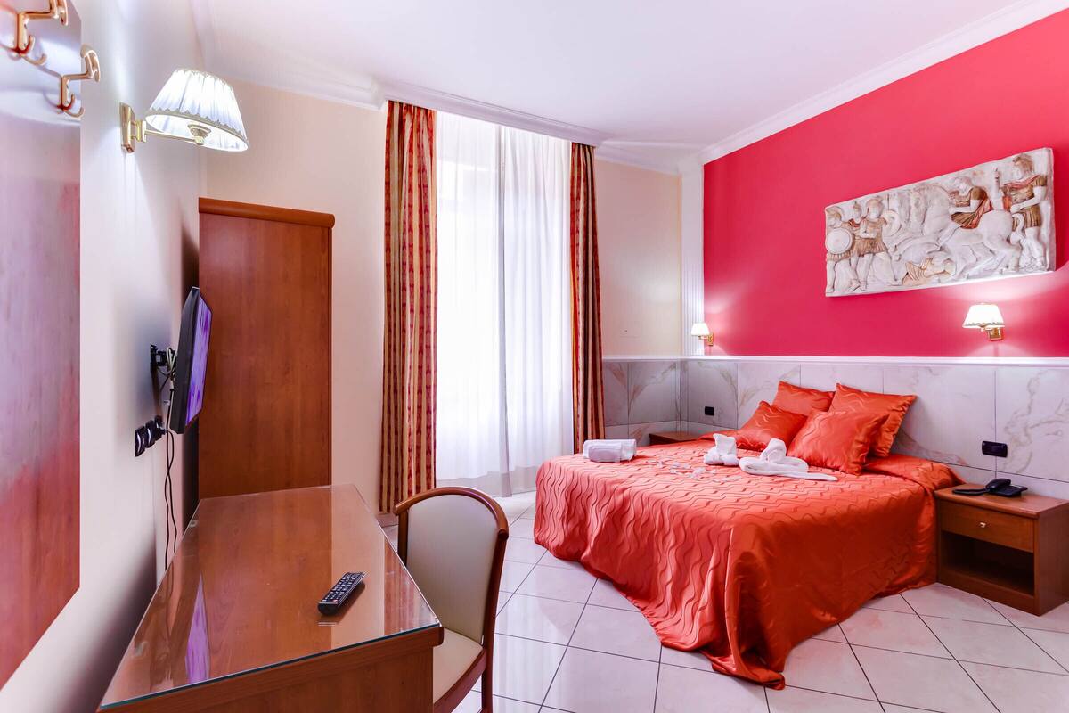 A hotel room with a bed, a desk, a chair, a TV, and a painting on the wall.