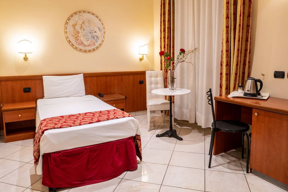 A hotel room with a bed, bedside table, desk, chair, and a small table with a vase of flowers.