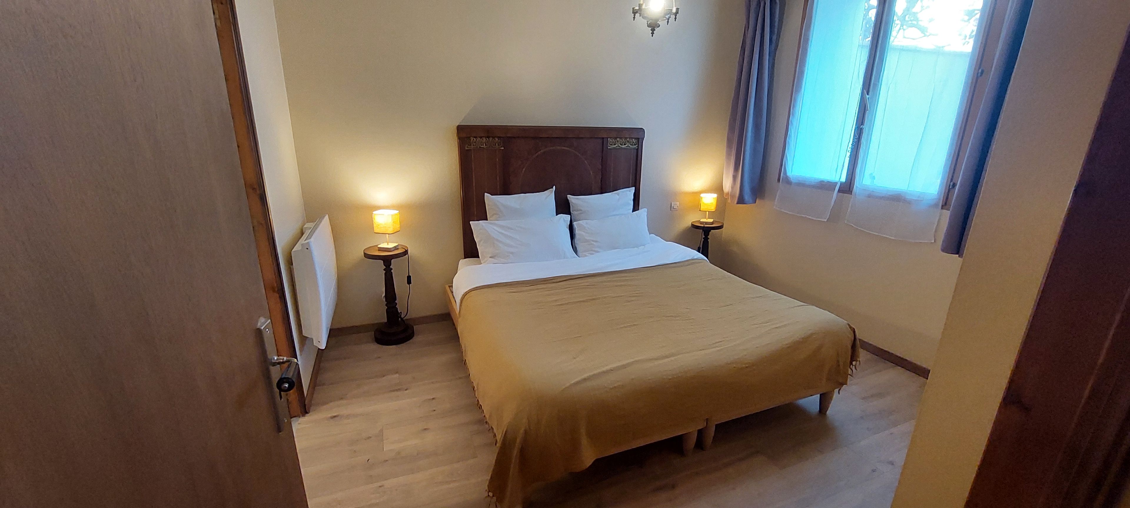 A hotel room with a bed, two bedside lamps, a window with curtains, and a radiator.