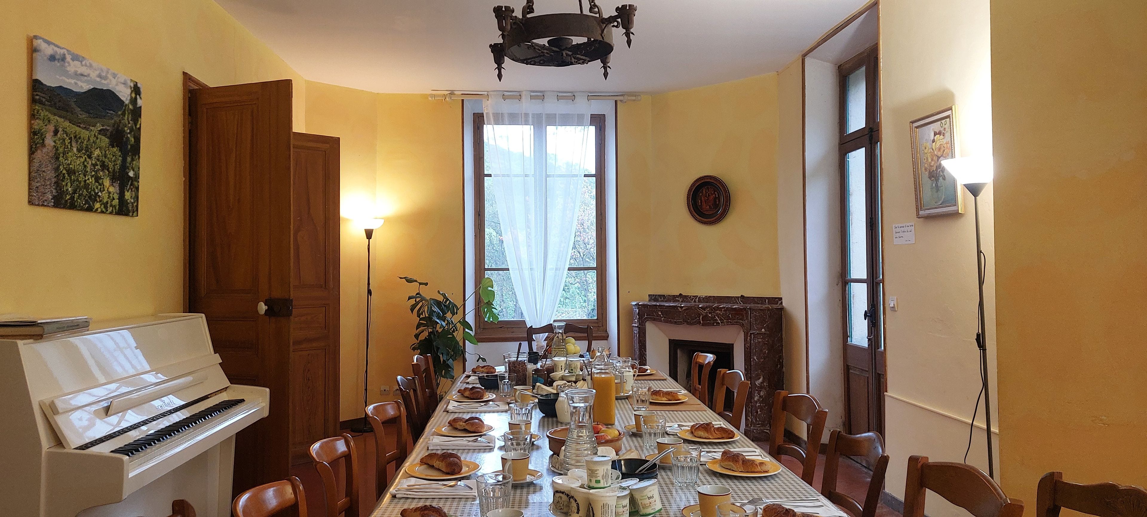Uma sala de jantar com um piano, uma mesa comprida posta para refeição e uma lareira.