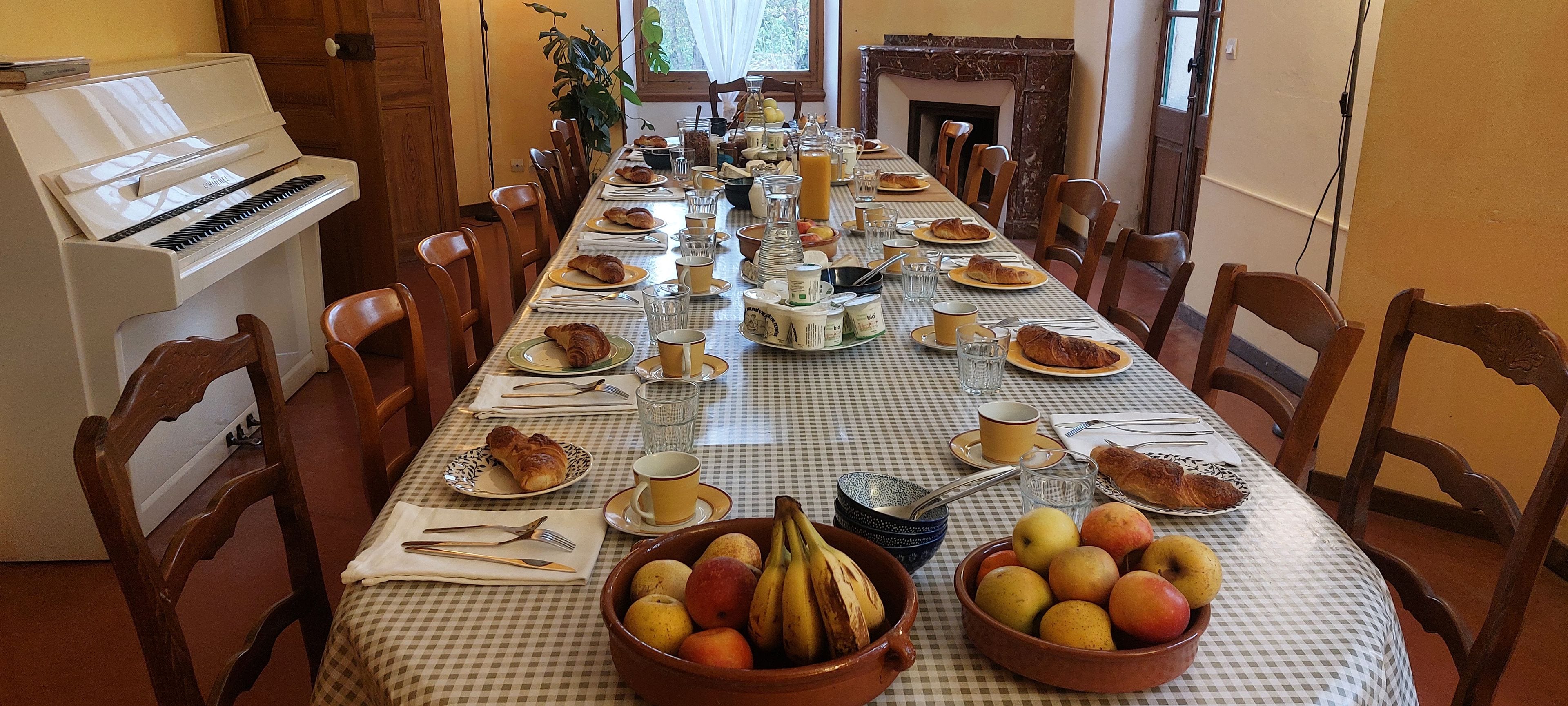 Una larga mesa de comedor dispuesta con platos, cubiertos y una variedad de alimentos, incluyendo frutas y pasteles, en una habitación con sillas de madera y un piano.