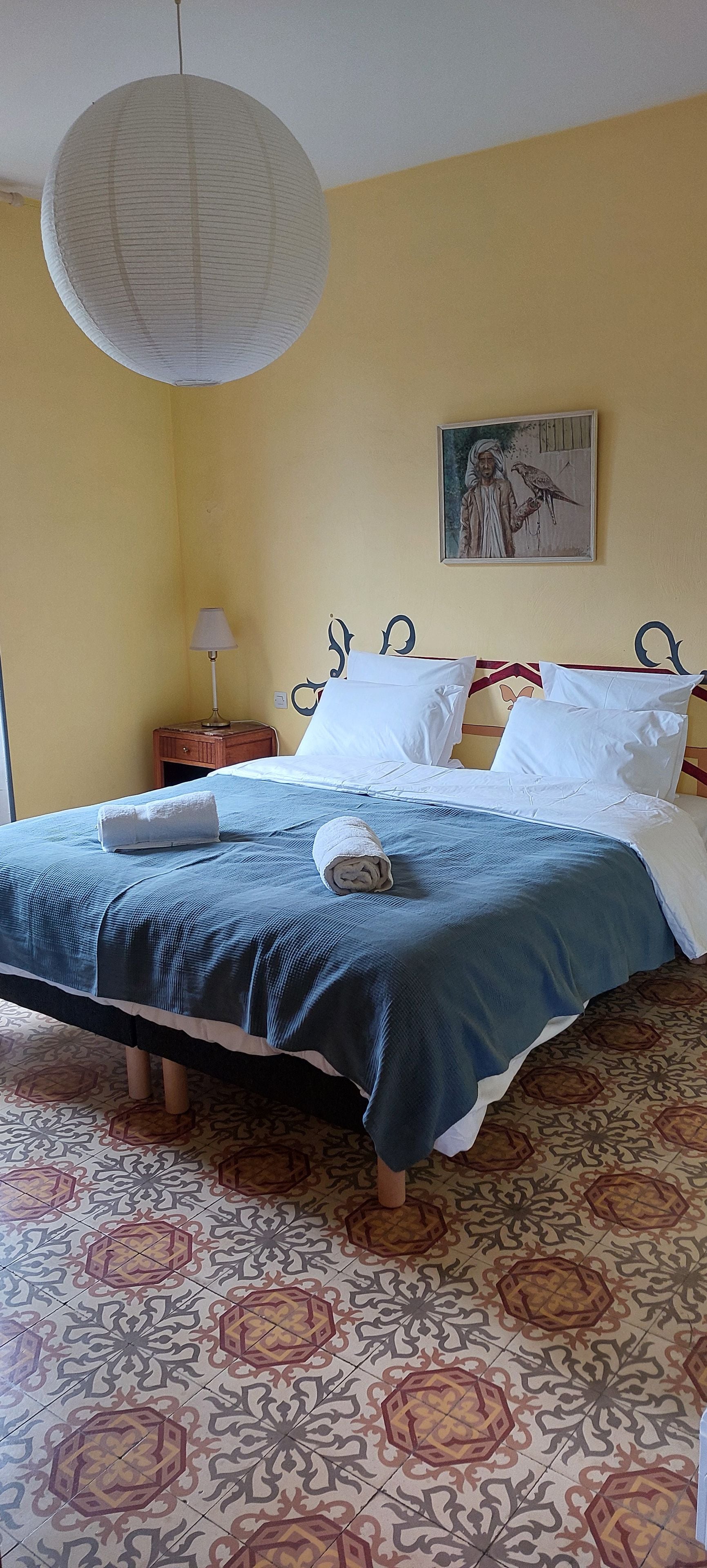 Un lit bien fait, recouvert d’une couvre-lit bleue et blanche, avec un oreiller blanc et une serviette enroulée posée sur le côté. On y trouve une table de chevet en bois avec une lampe, un tableau accroché au mur et un sol à motifs.