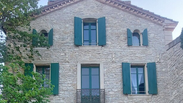 A stone building with a central entrance, green shutters, and a small balcony.