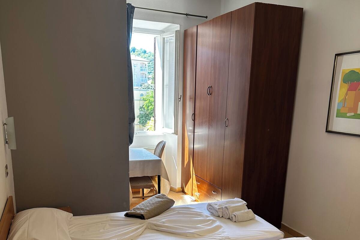 Ein Hotelzimmer mit zwei Betten, einem Holzschrank, einer Essecke und einem Fenster mit Tageslicht.