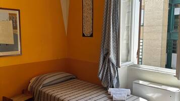 A single bed with striped bedding, a small bedside table, a radiator, and a window with curtains.