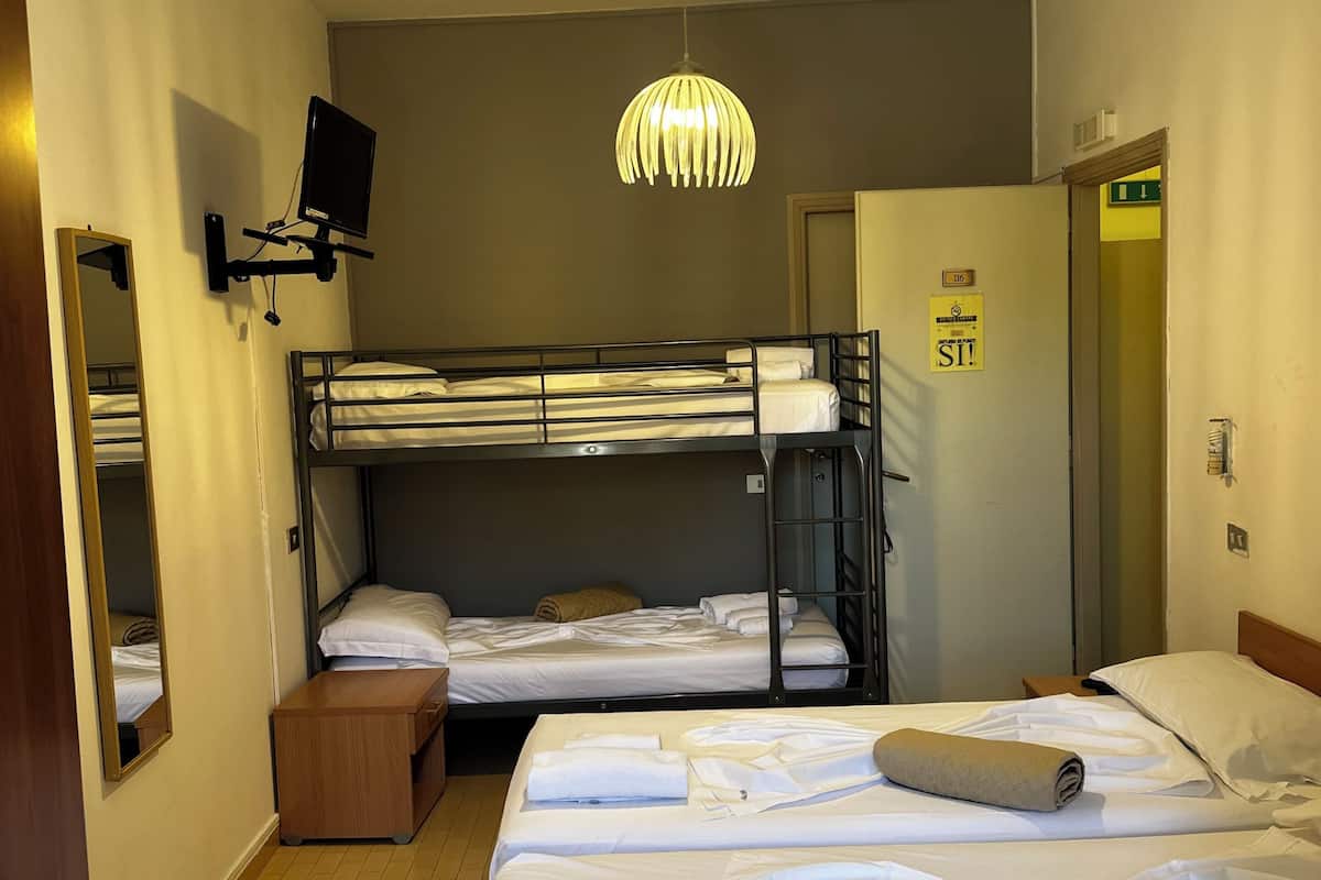 Ein Hotelzimmer mit Etagenbetten, einem Doppelbett, einem an der Wand befestigten Fernseher, einer Hängeleuchte und einer Tür mit Fluchtzeichen.
