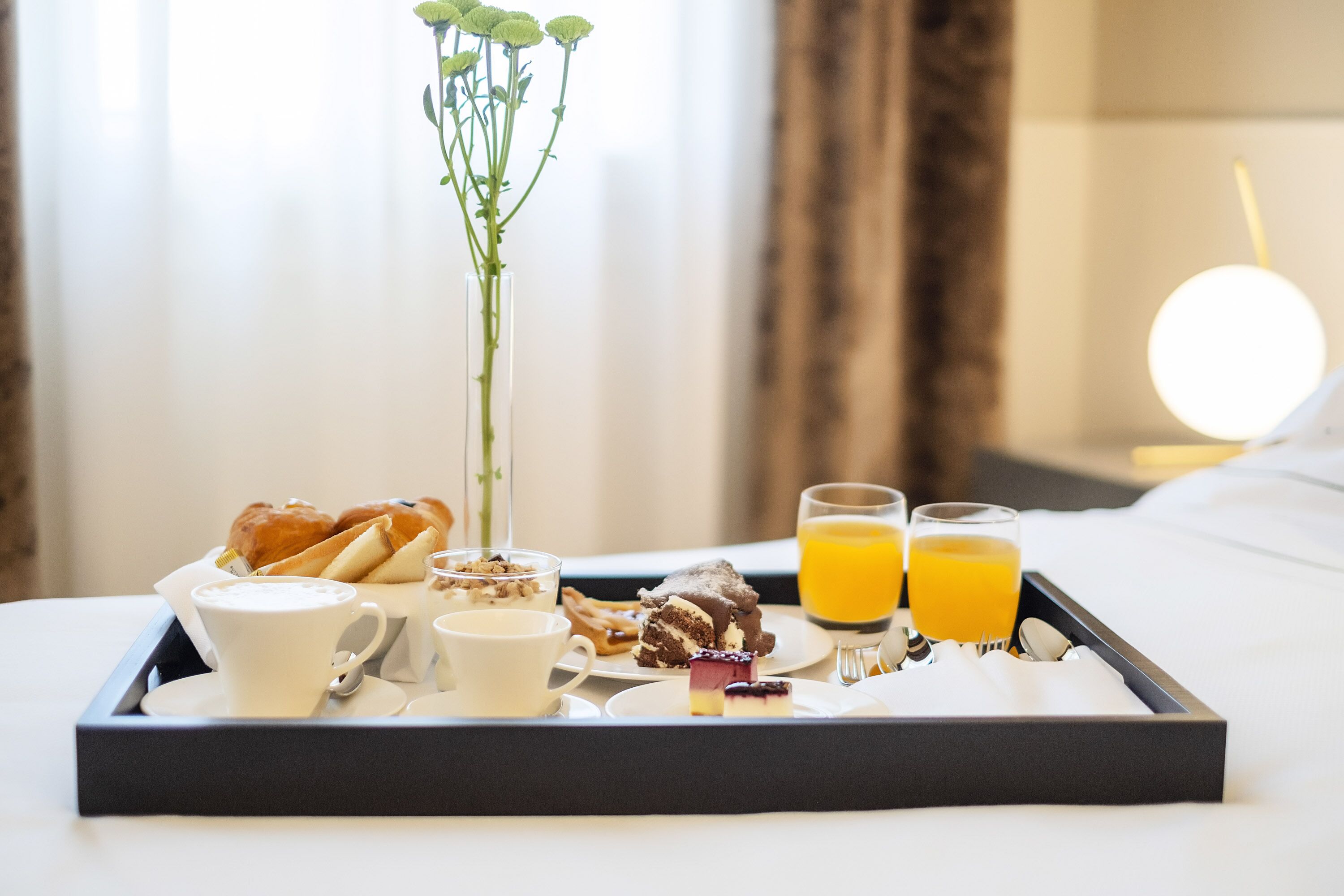 A breakfast tray with croissants, a coffee cup, a small cake, and orange juice on a bed.