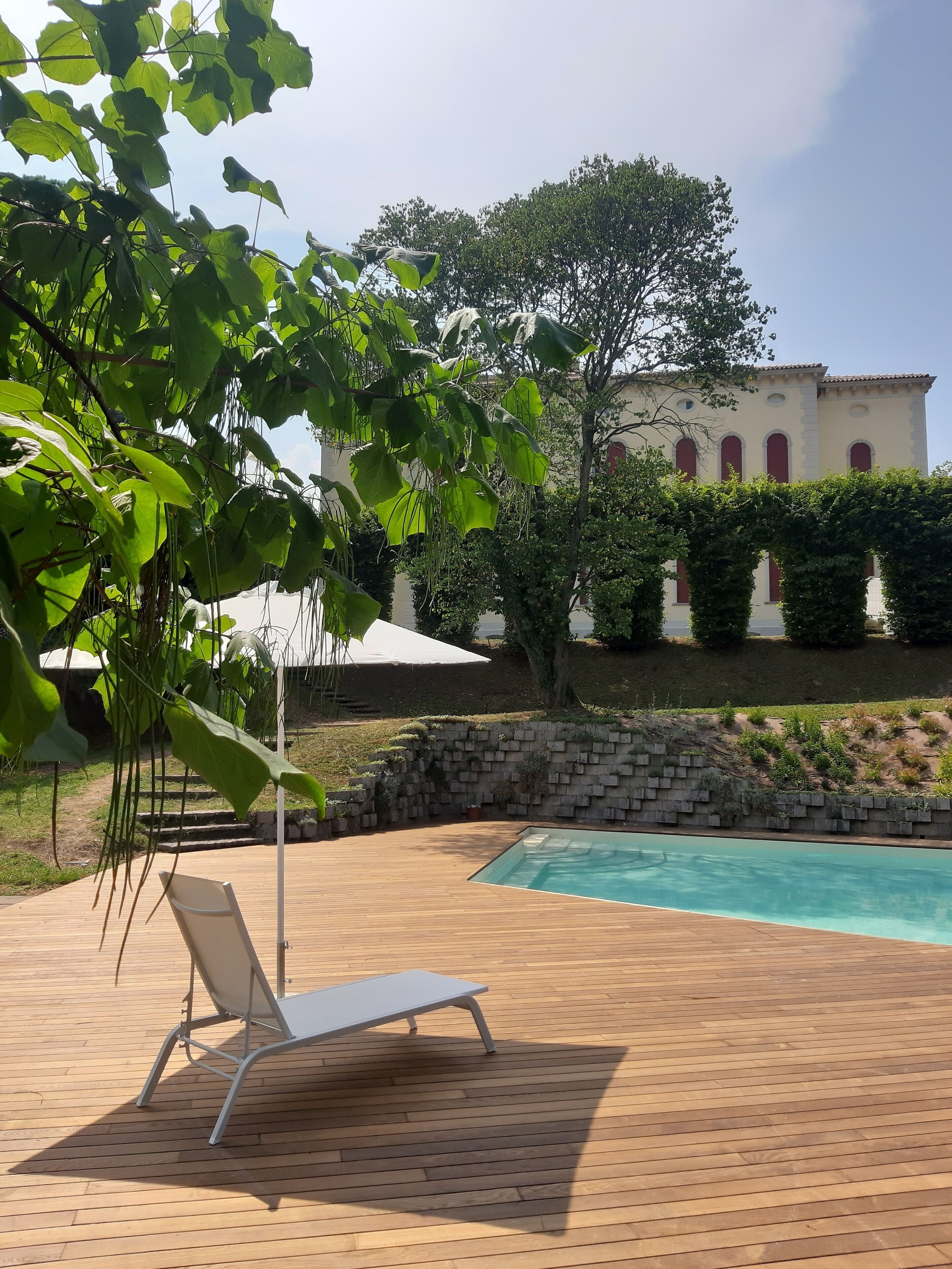 Área de piscina com espreguiçadeira, um pátio de tijolos e um edifício com janelas em arco ao fundo.
