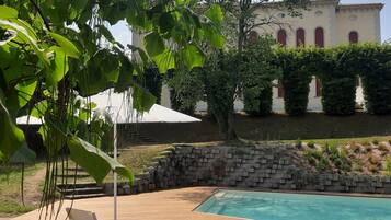Área de piscina com espreguiçadeira, um pátio de tijolos e um edifício com janelas em arco ao fundo.