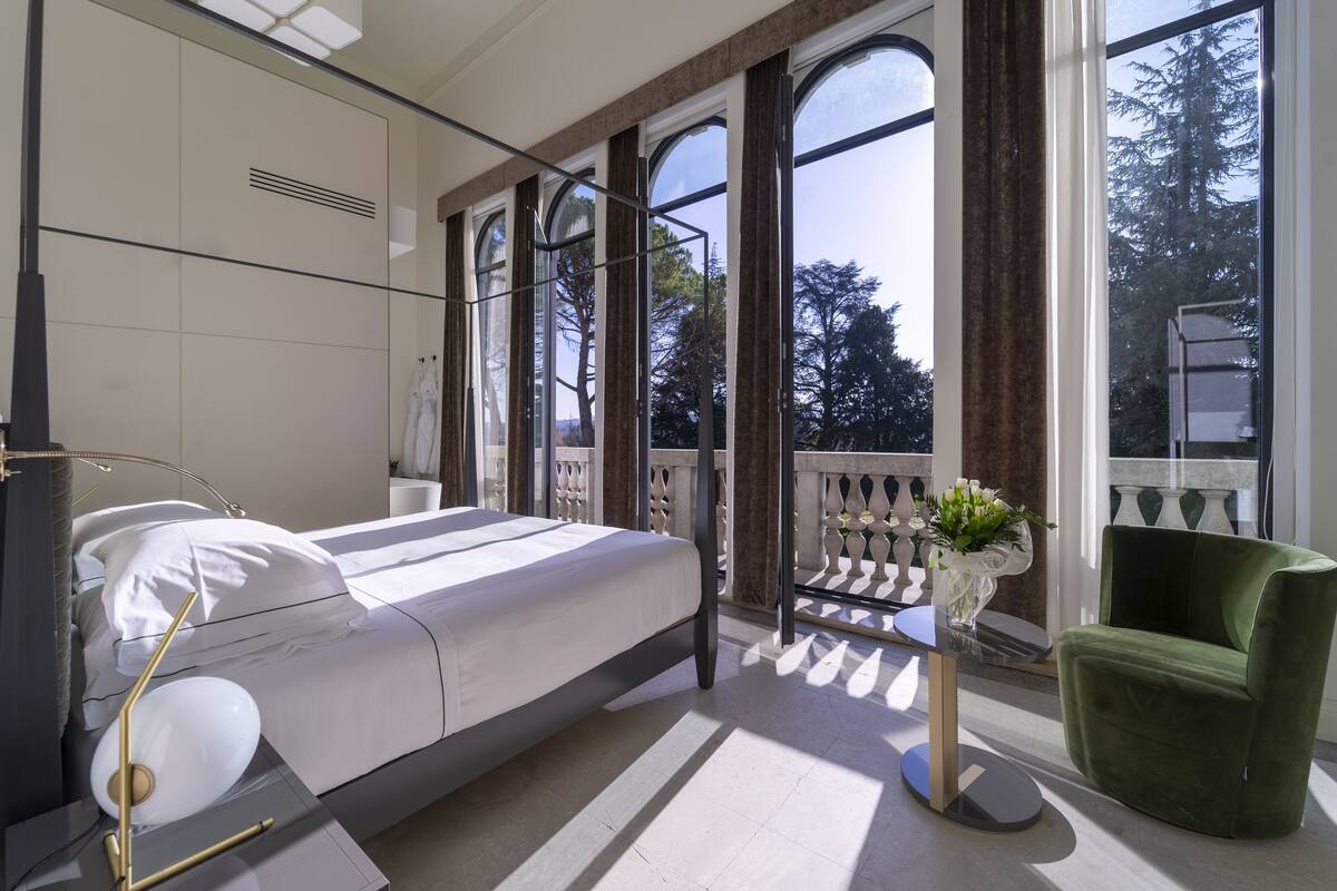 A modern bedroom with a bed, a green armchair, a small table with a vase of flowers, and large windows with a view of trees.