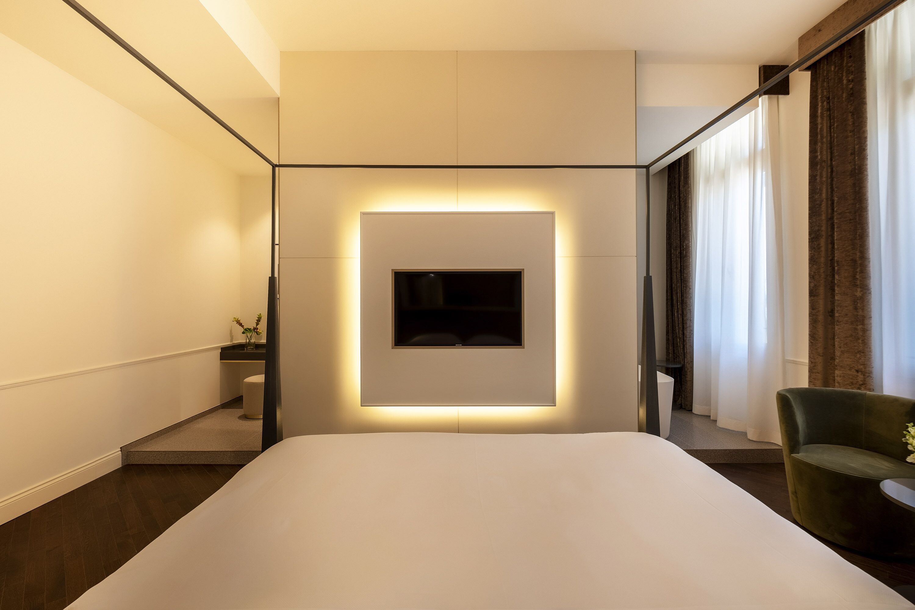 A modern hotel room with a large bed, a mounted TV, a small seating area, and a window with curtains.