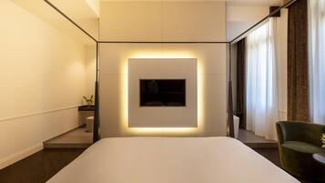 Una habitación de hotel moderna con una cama grande, un televisor montado, un pequeño rincón de descanso y una ventana con cortinas.