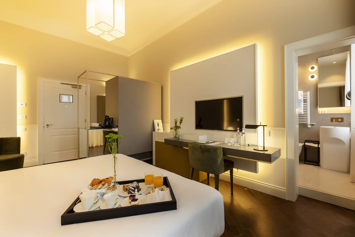 A modern hotel room with a large bed, a desk with a flat-screen TV, a dining area with breakfast, and a bathroom visible through an open door.