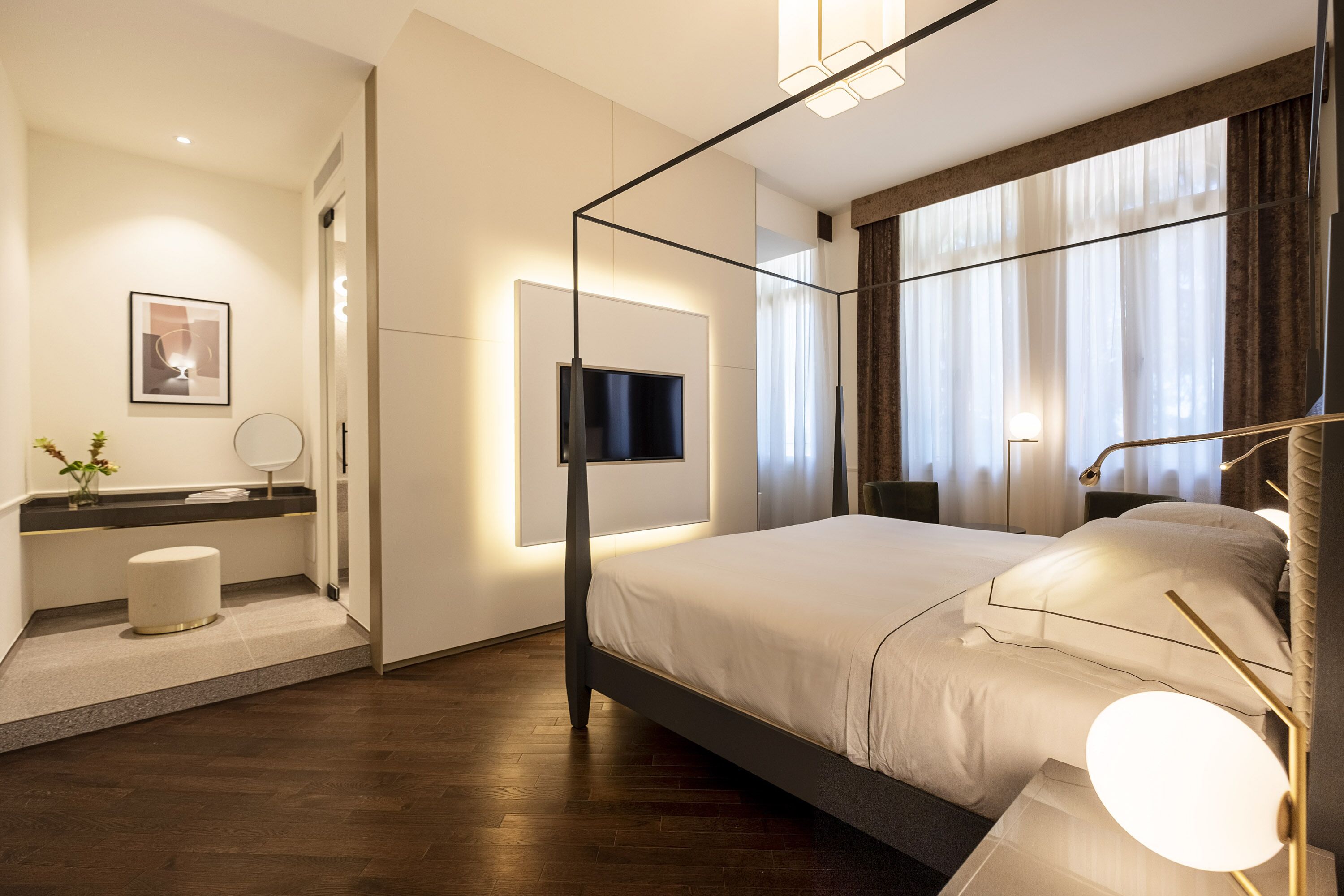 Habitació d'hotel moderna amb un llit gran, una televisió plana penjada a la paret, una tauleta de nit amb una làmpada i un mirall rodó petit.