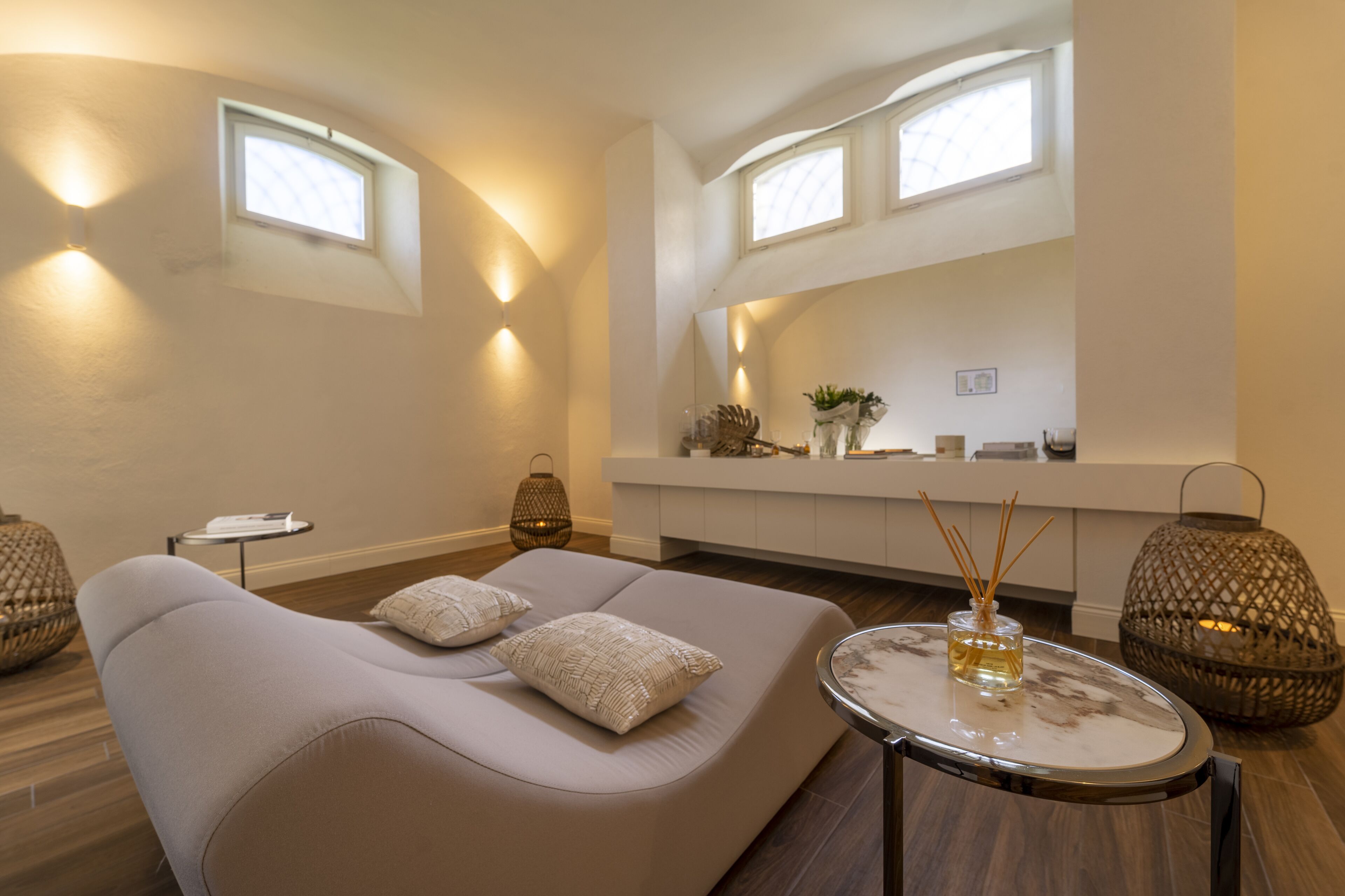 A modern living room with a sofa, a round table, and decorative items. There are two windows providing natural light.