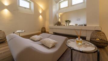 A modern living room with a sofa, a round table, and decorative items. There are two windows providing natural light.