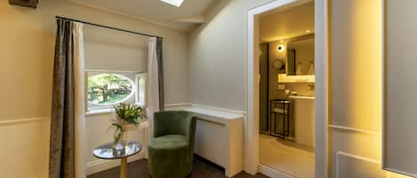 Una habitación con tragaluz, una silla verde, una mesita con una planta y vistas al baño.