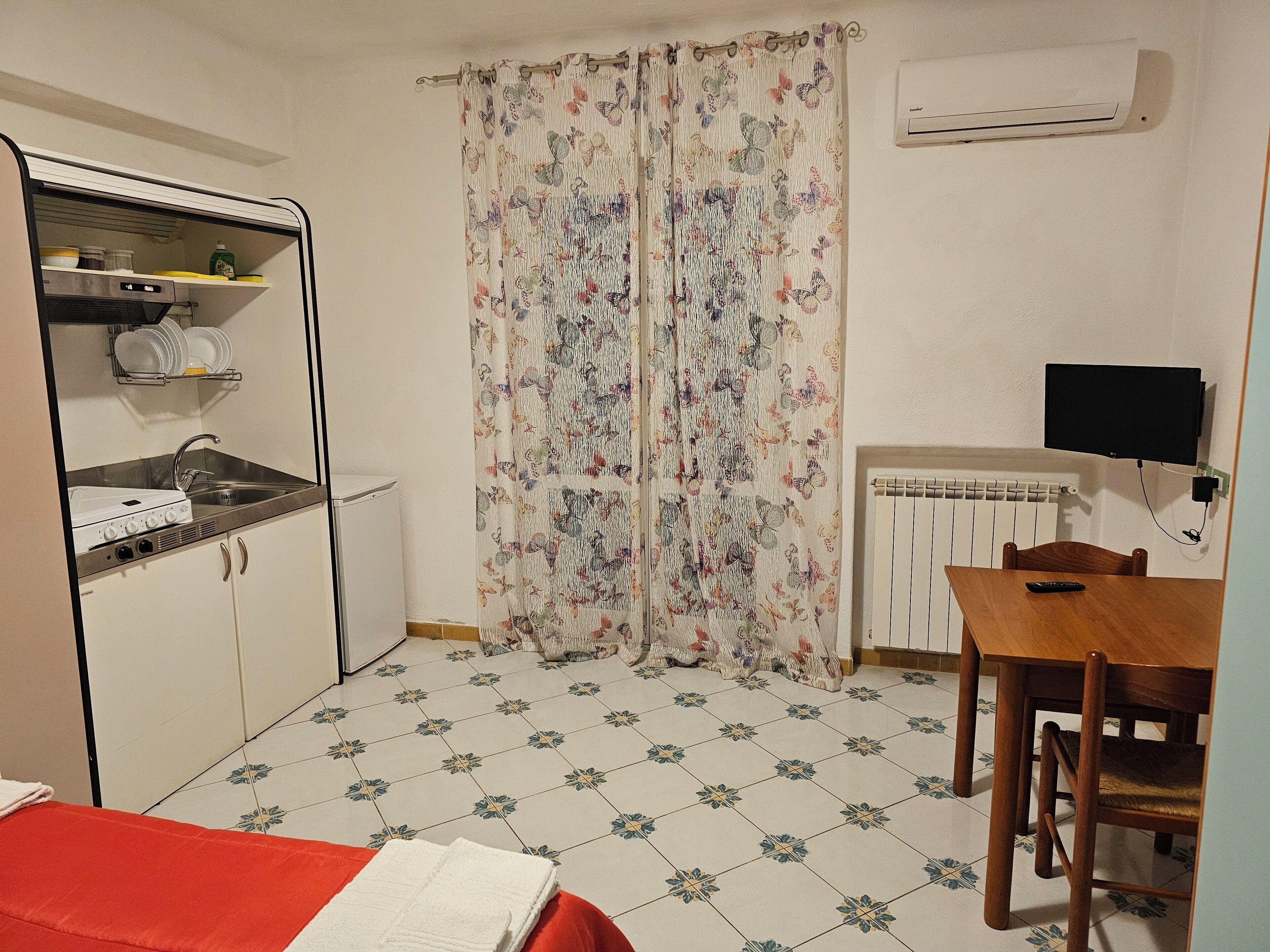 A compact kitchen with a built-in stove, a sink, and a small refrigerator. There is a dining table with chairs and a television mounted on the wall.