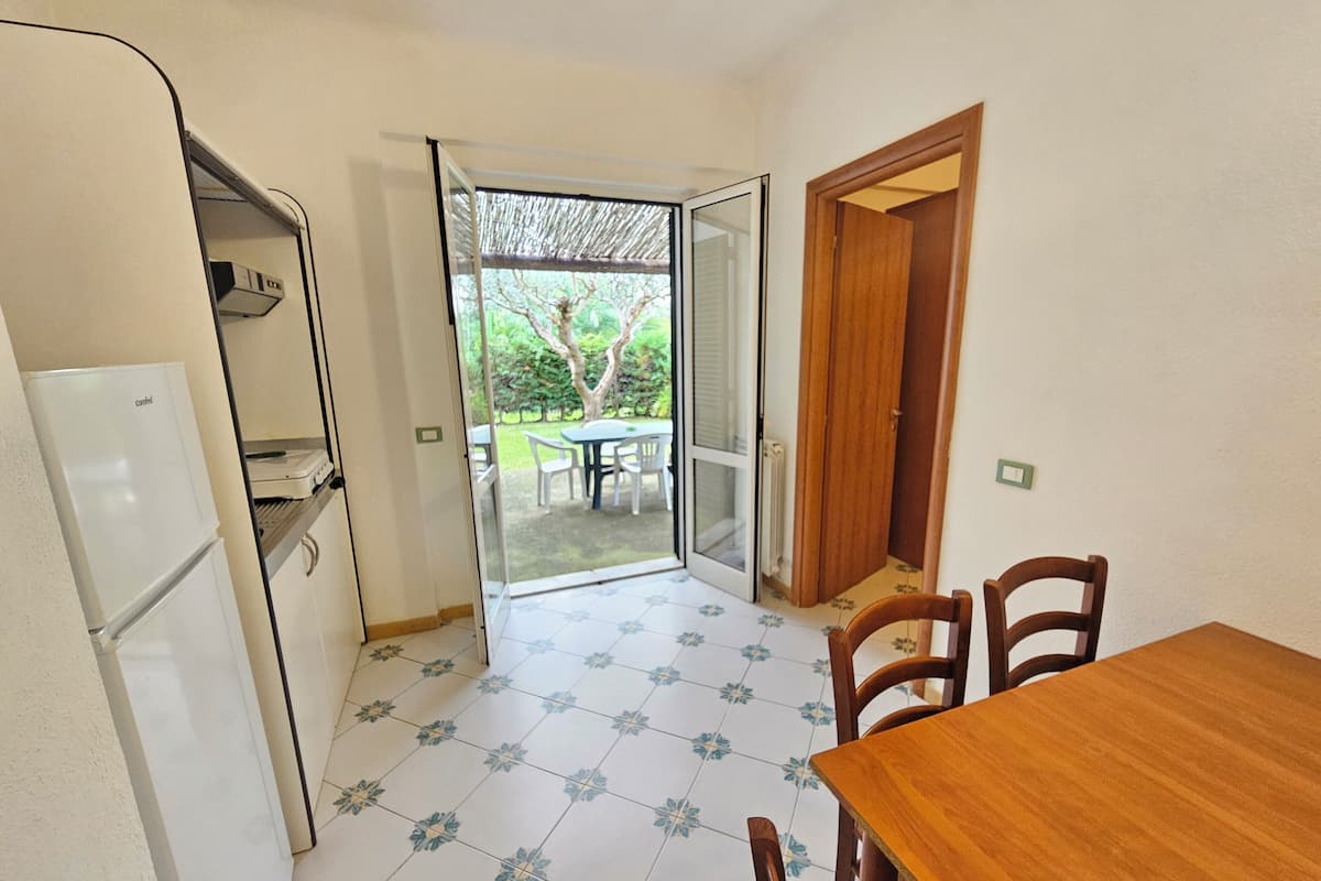 Una cocina compacta con refrigerador, una mesa de comedor con sillas y un ventanal corredero que da a un patio exterior con mesa y sillas.