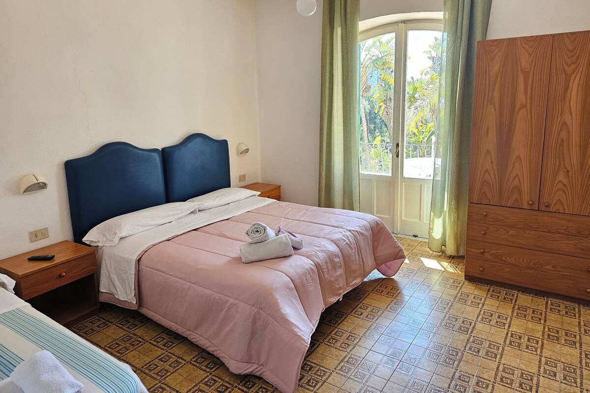 Un dormitorio con cama, mesitas de noche, armario y ventana con cortinas.