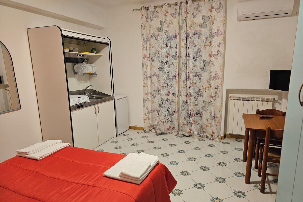 Una habitación compacta con cama, una kitchenette pequeña, un comedor y una ventana con cortinas florales.