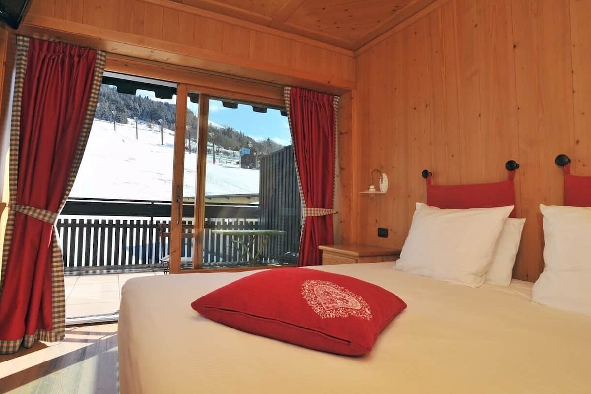 A wooden cabin bedroom with a large bed, red curtains, and a view of a snowy landscape.