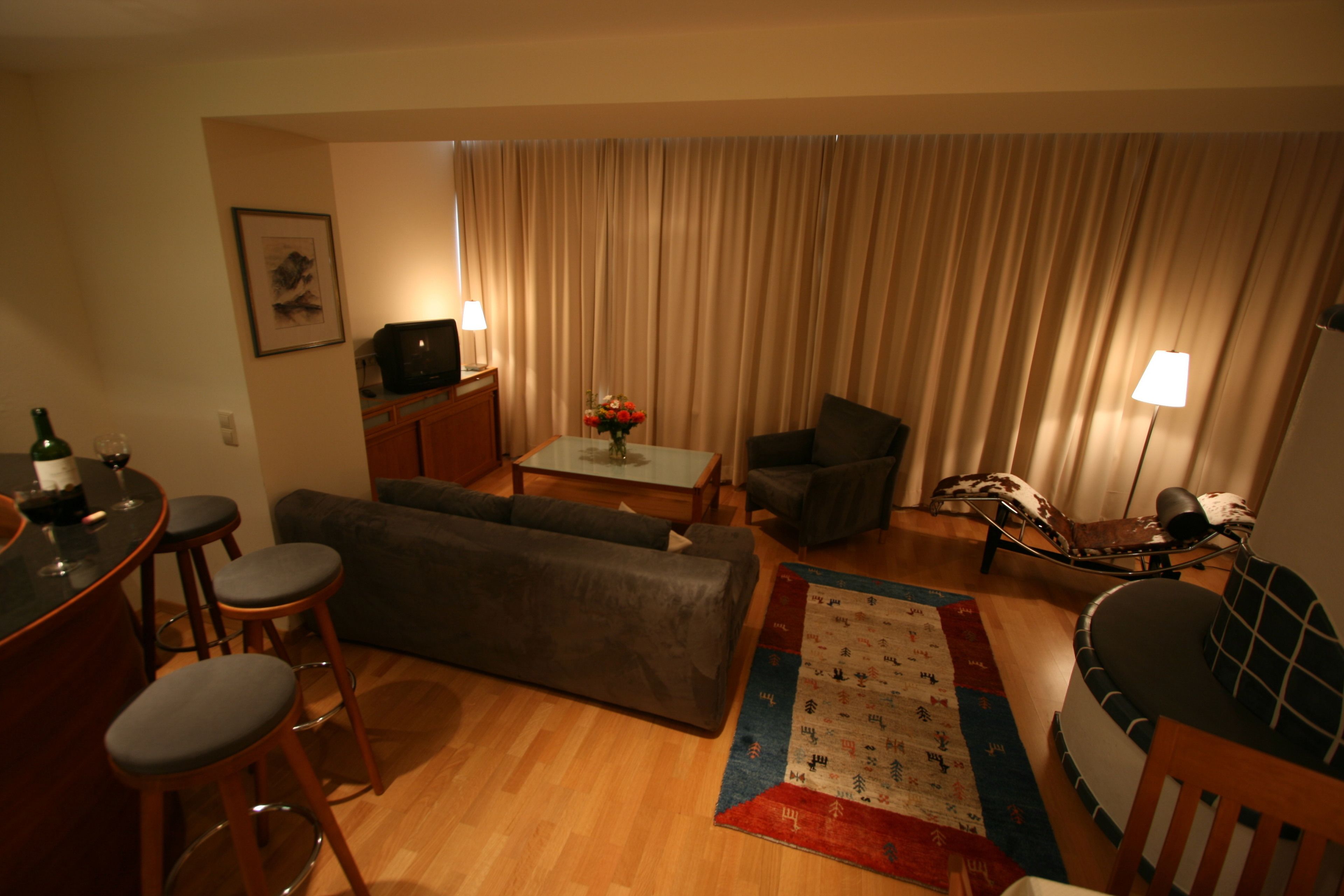 Ein Wohnzimmer mit einer Couch, zwei Sesseln, einem Couchtisch und einem Fernseher. Es gibt Barhocker, eine Lampe und einen Teppich.