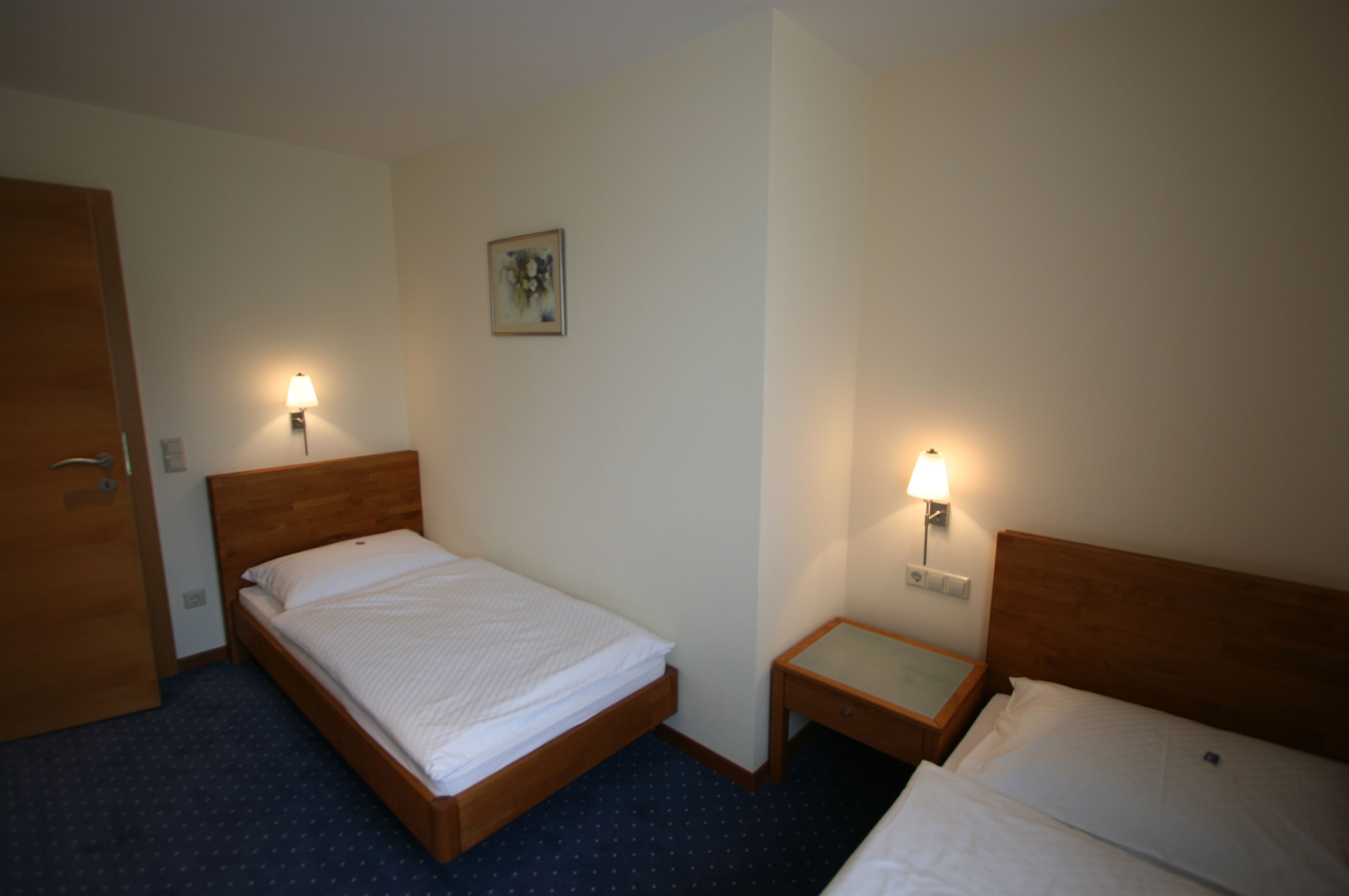 Une chambre d’hôtel avec deux lits simples, une tête de lit en bois, une table de chevet avec une lampe, une petite table et un tableau au mur.