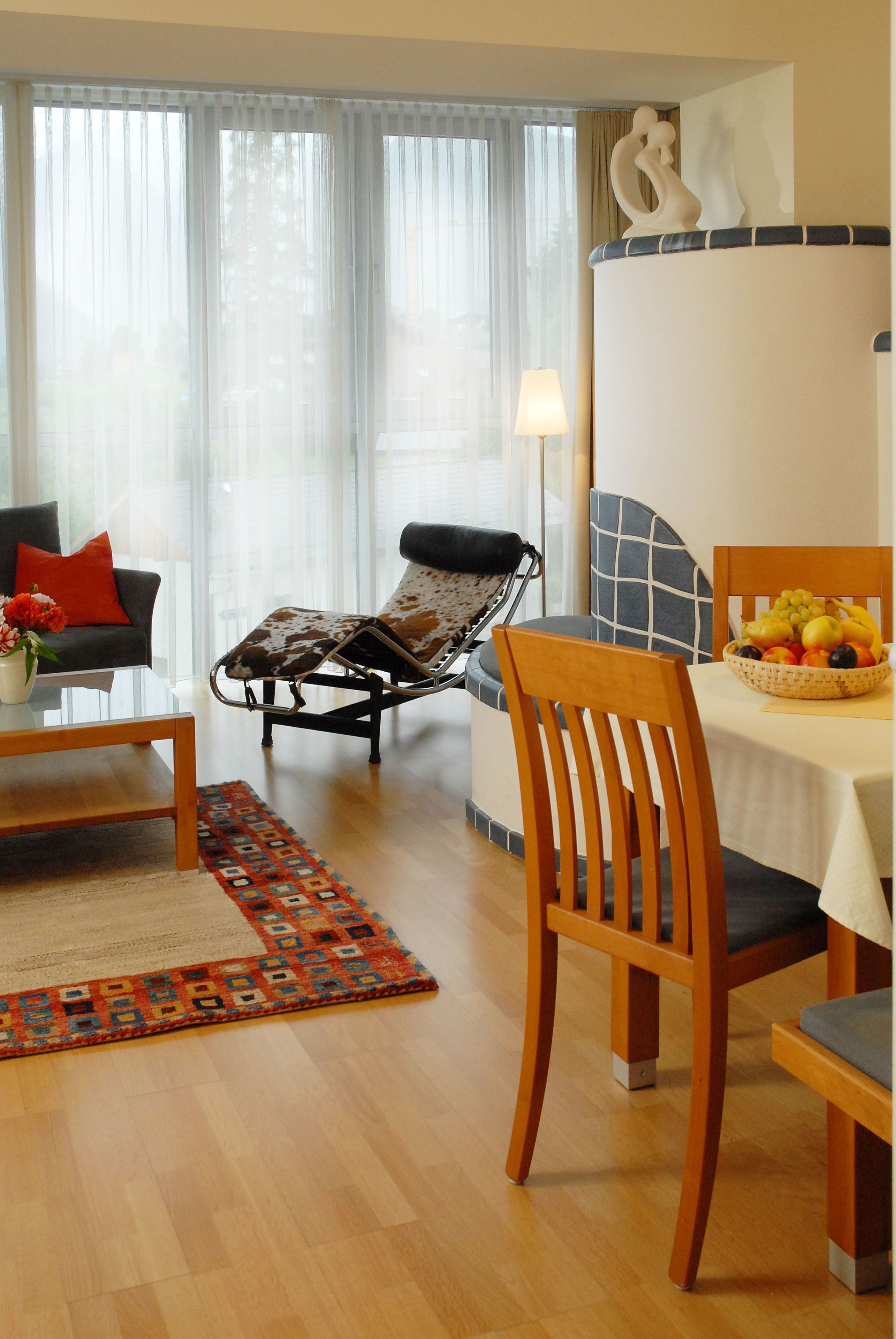 A living room with a sofa, a coffee table, a dining table with chairs, a patterned rug, and a window with curtains.