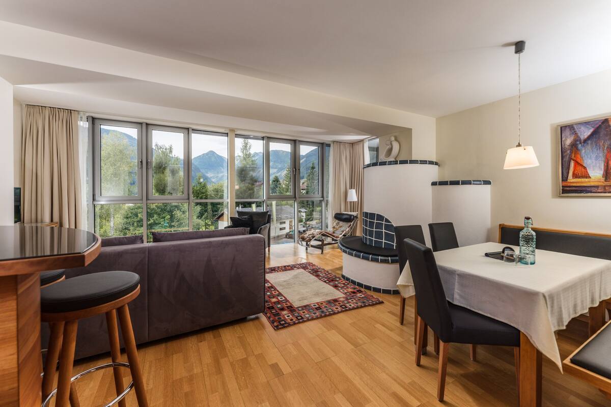 A modern living room with a sofa, dining table, and bar stools. Large windows offer a view of mountains.