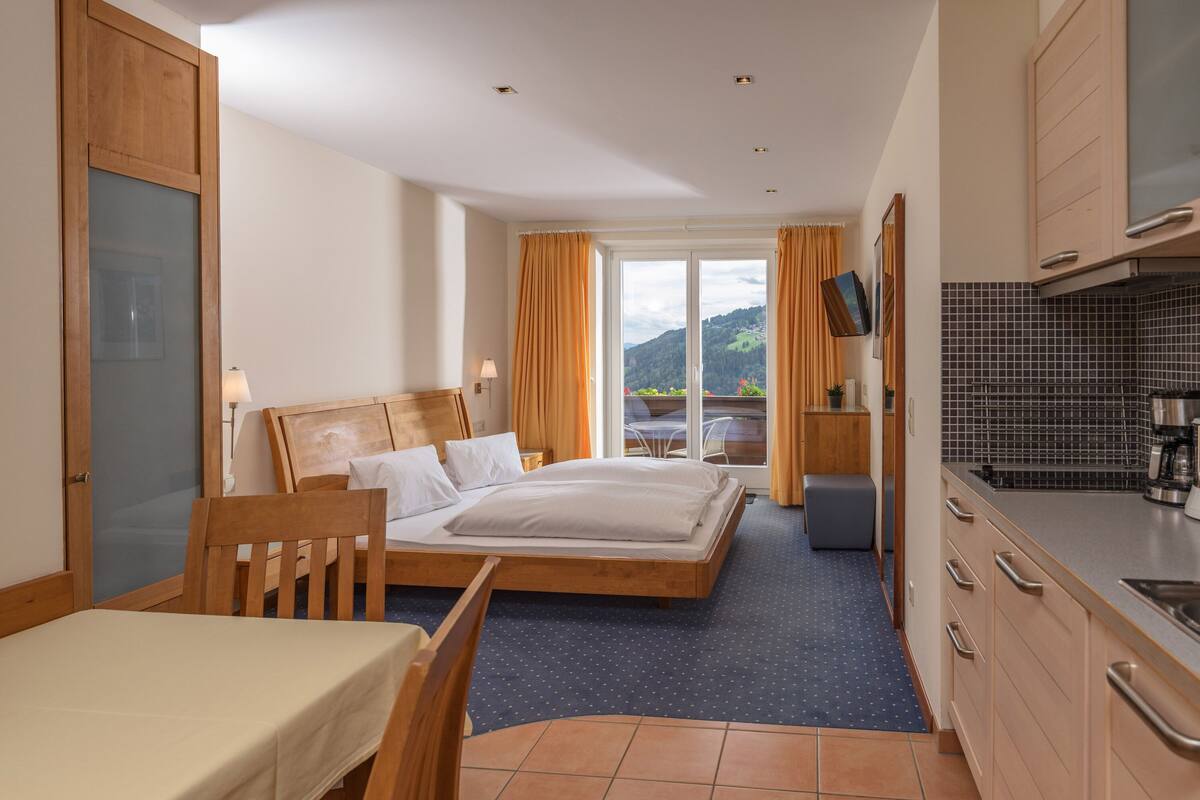 A hotel room with a bed, a desk, a chair, a small table, a kitchen area, and a view of the outdoors.