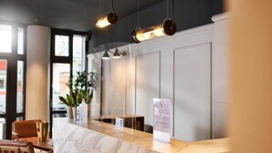 A hotel reception area with a marble counter, hanging pendant lights, a potted plant, and a signboard. - Hôtel Les Negociants (Valence)