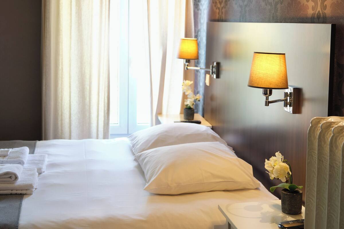 A hotel room with a neatly made bed, two bedside lamps, a small table with a vase of flowers, and a chair.
