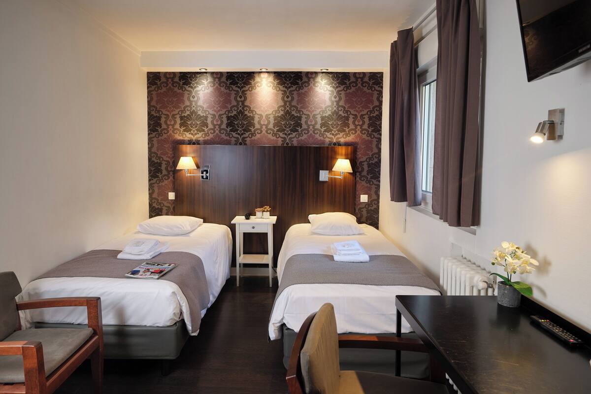 A hotel room with two single beds, a small table, a chair, a desk, and a TV.