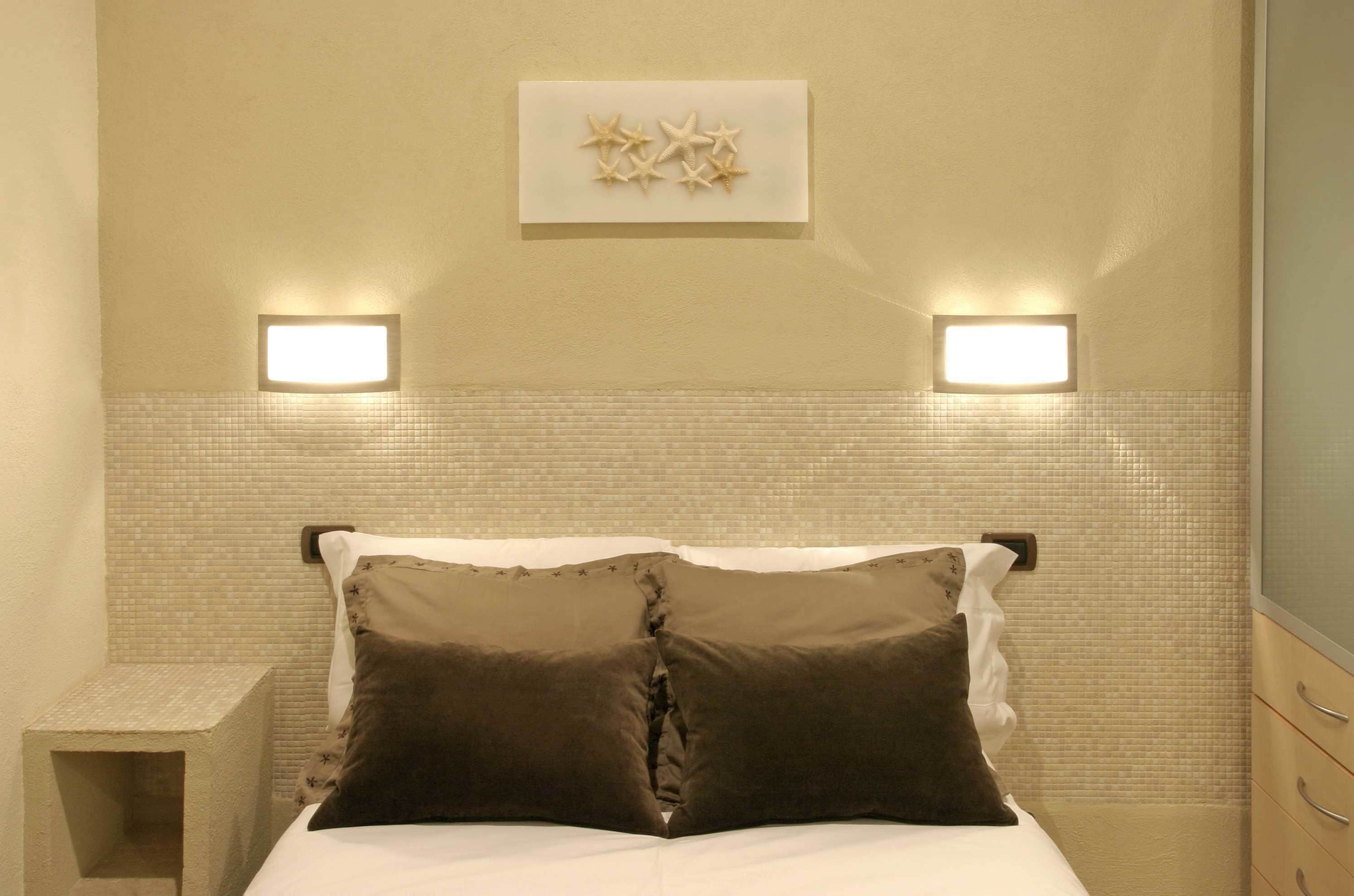 A hotel room with a bed, two bedside lamps, a wall-mounted light fixture, and a small shelf.