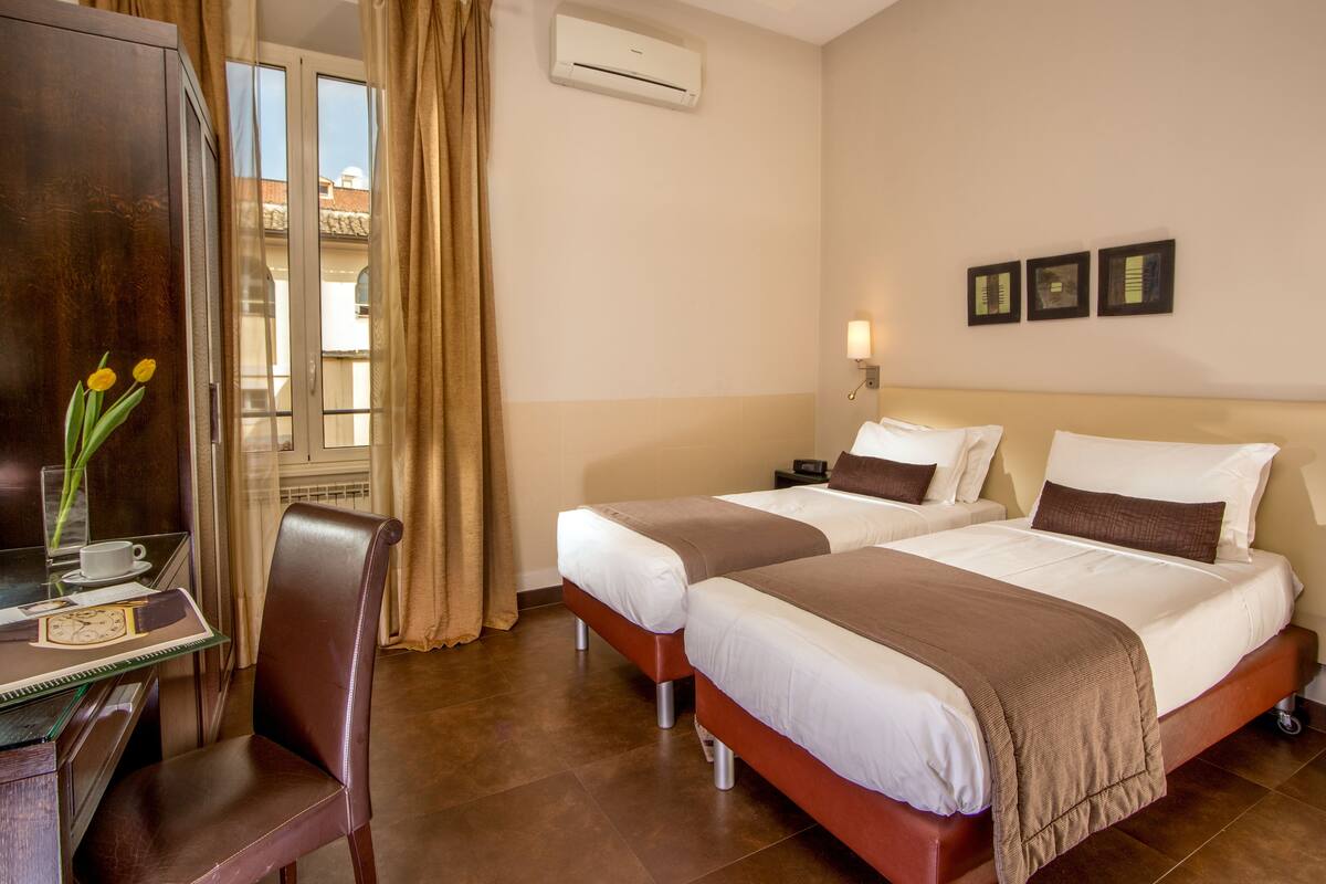 Habitación de hotel con dos camas, un escritorio con silla, un florero con flores y una ventana con cortinas.