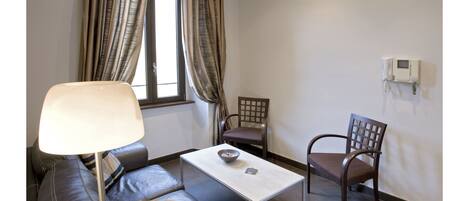 A modern living room with a sofa, a table, and two chairs. There is a window with curtains, a lamp, and a wall-mounted thermostat.