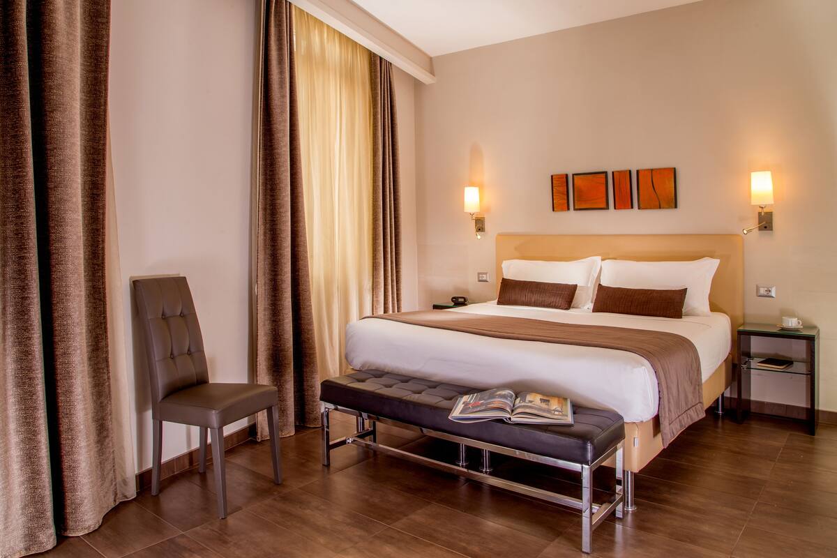 Una habitación de hotel con una cama grande, una mesita de noche, una silla y un ventanal con cortinas.