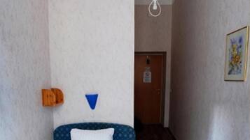 A small, narrow room with a bed, a wooden floor, a white ceiling, and a door at the end.