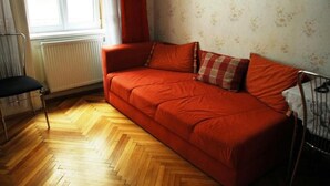 Una habitación con suelo de madera en patrón espiga, un sofá rojo, un radiador, una ventana con cortinas blancas y un cuadro enmarcado en la pared.