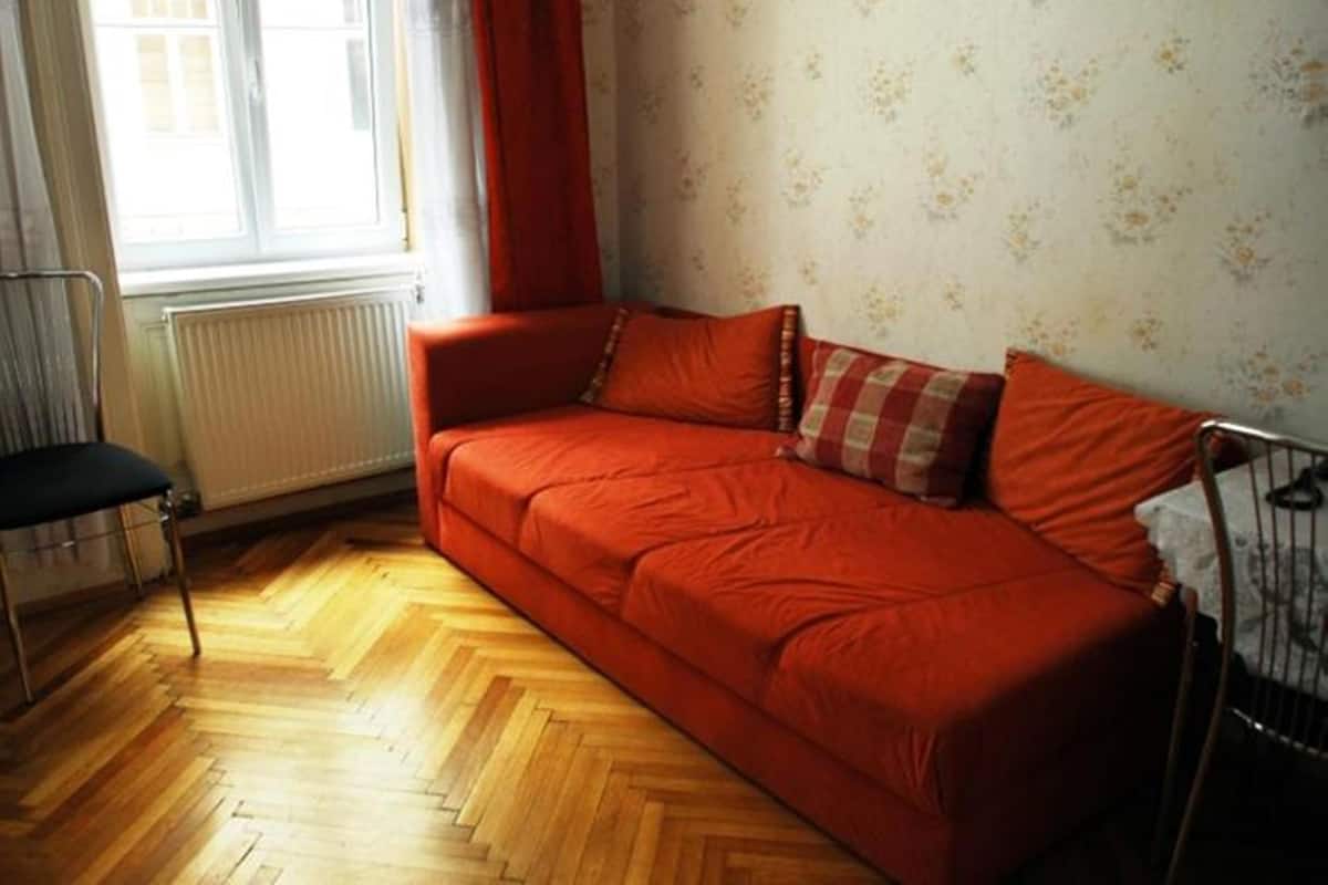 Um quarto com piso de madeira em padrão herringbone, um sofá vermelho, um radiador, uma janela com cortinas brancas e uma moldura com um quadro na parede.