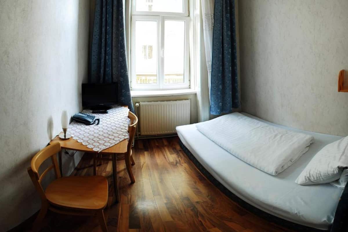 Um quarto pequeno com uma cama, uma escrivaninha com cadeira, uma televisão e uma janela com cortinas.