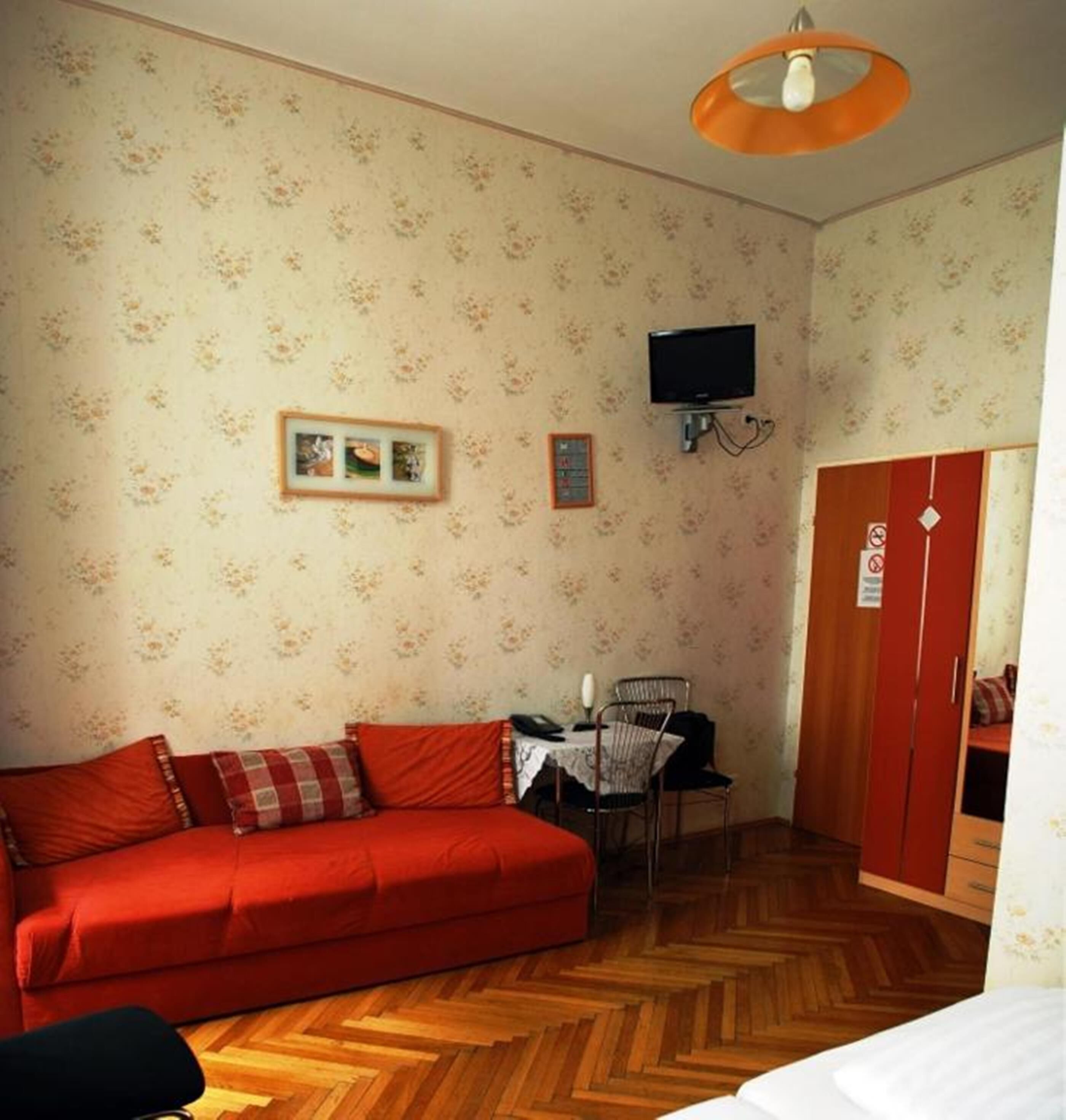 Quarto com sofá vermelho, uma mesa pequena com cadeiras, televisão fixada na parede e um guarda-roupa.