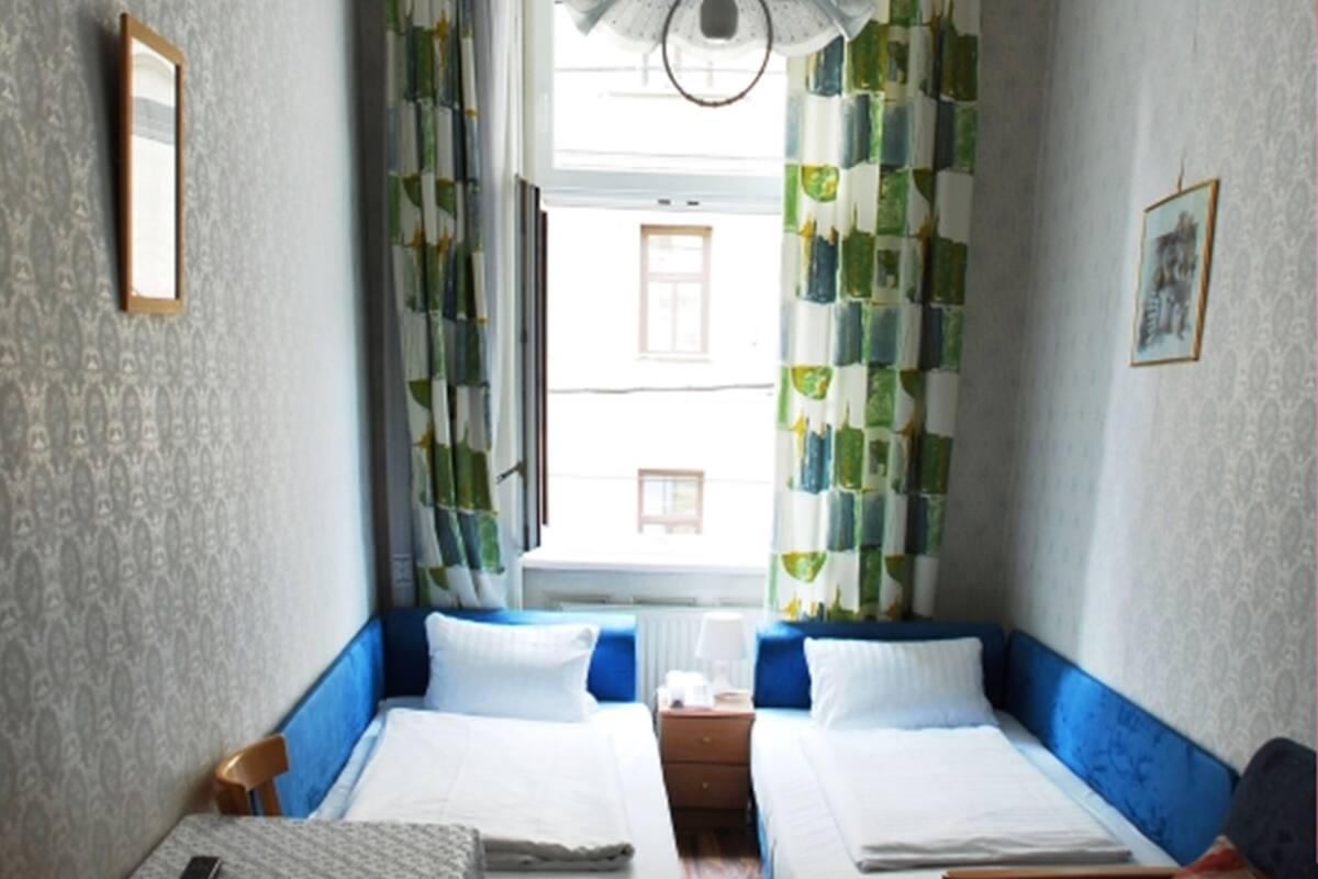 Quarto pequeno e acolhedor com duas camas de solteiro, uma mesa com abajur e uma janela com cortinas.