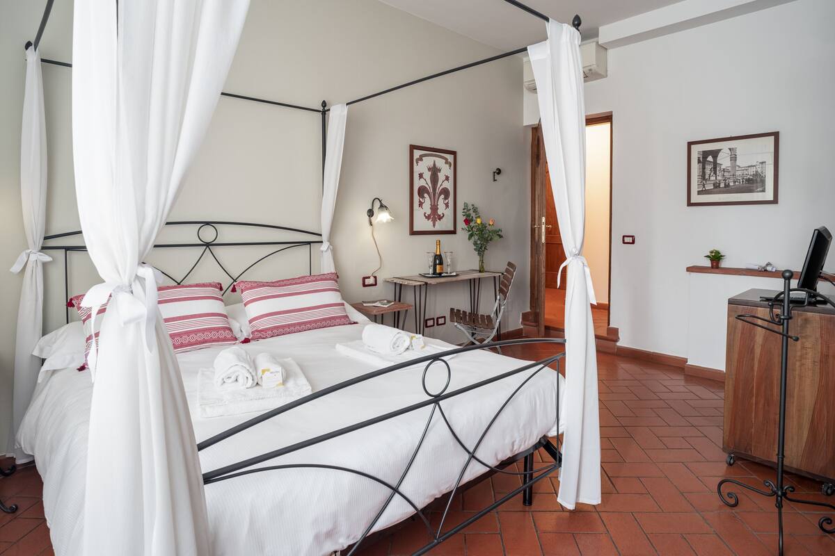 A four-poster bed with white bedding and red pillows, a small table with a bottle and flowers, and a desk with a computer.