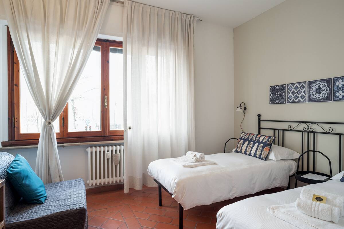 A bedroom with two beds, a window with curtains, a radiator, and a wall with decorative tiles.